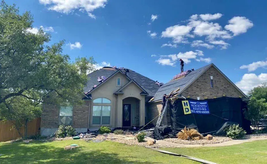 House with roof being repaired under a blue sky; workers on roof with tarp.