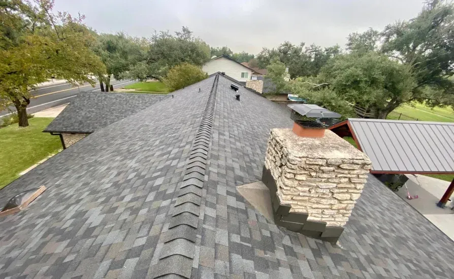 A house roof with gray shingles, a stone chimney, and trees in the background under a cloudy sky.