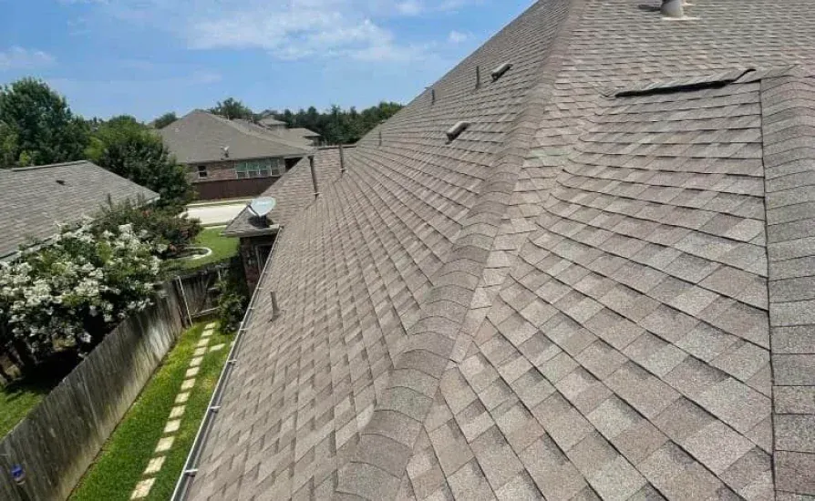 Brown shingle roof on a house with a blue sky and other houses in the background.
