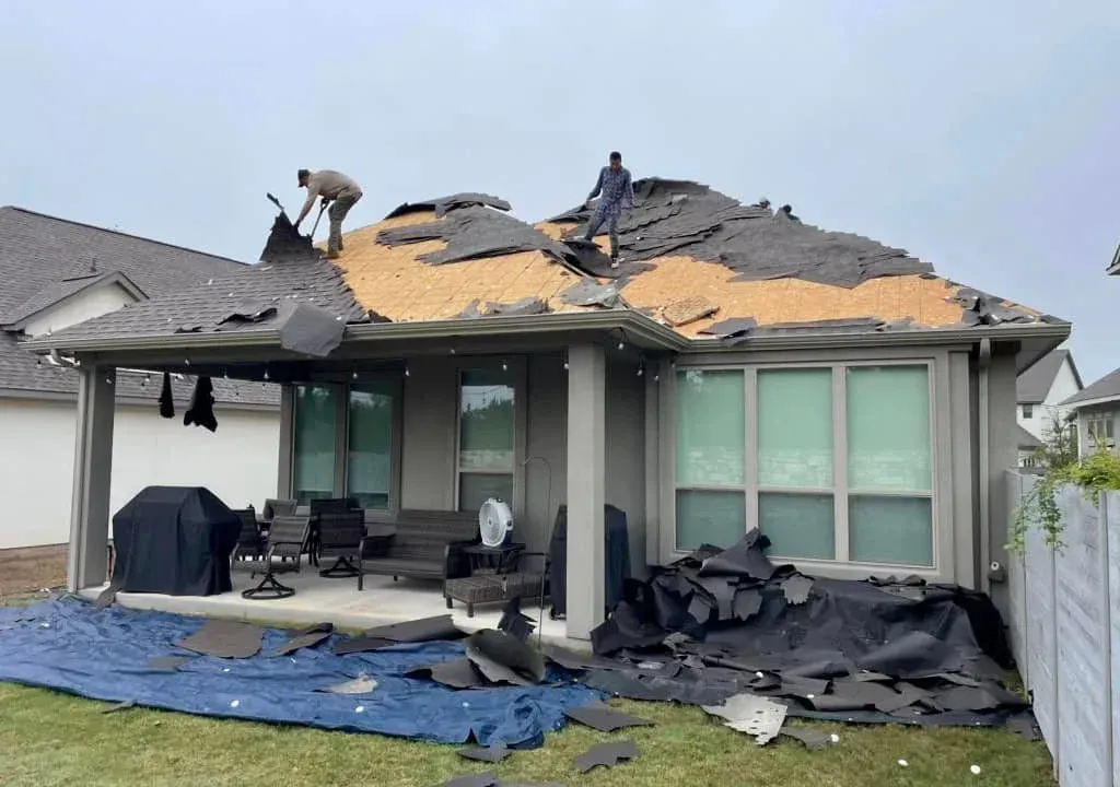Two workers removing shingles from a house roof, debris on a blue tarp below, overcast sky.