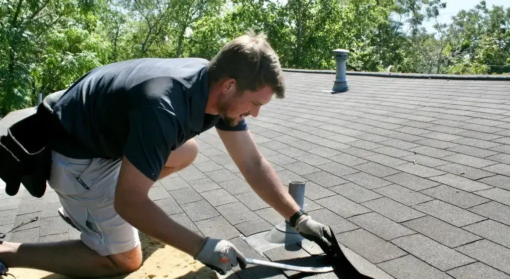 Roofer kneeling on a rooftop, repairing shingles. Wearing gloves and a tool belt, working in daylight.