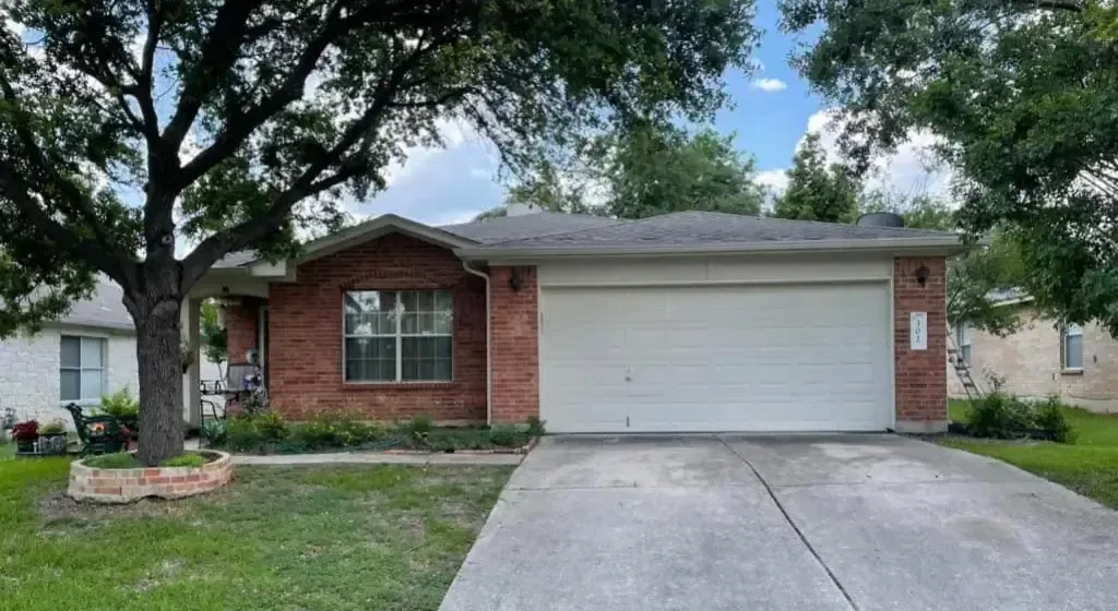 Red brick house with a two-car garage, under a tree, and a concrete driveway.