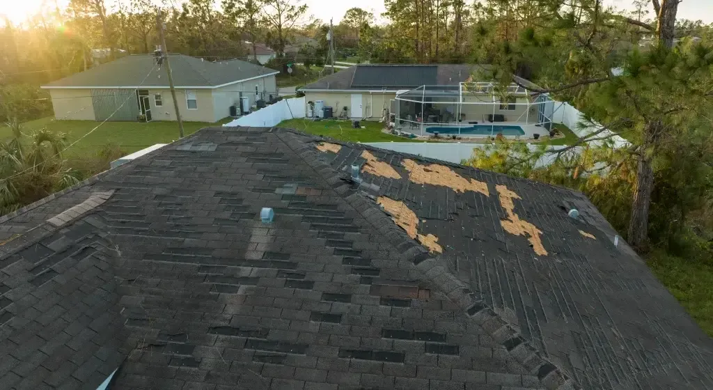 Damaged roof with missing shingles on a residential house after a storm. Homes and trees are in the background.