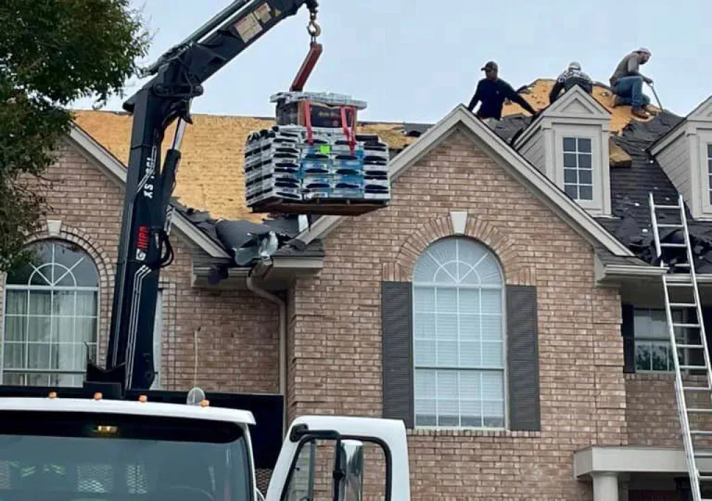 Crane lifting shingles to a roof where workers are replacing the roofing on a brick house.