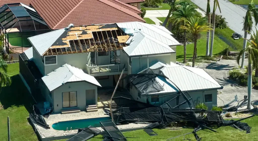 House with significant roof and structure damage, pool in foreground, palm trees in background.