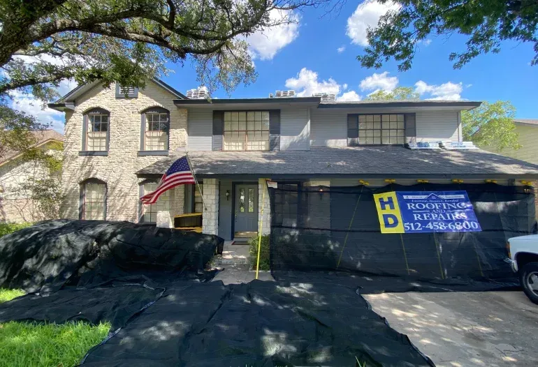 House with roofing work in progress, tarp covers, American flag, blue sky.