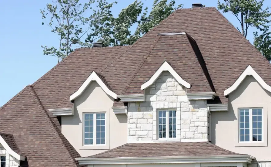 Brown shingled roof on a two-story house with white trim, stone accents, and windows against a blue sky.
