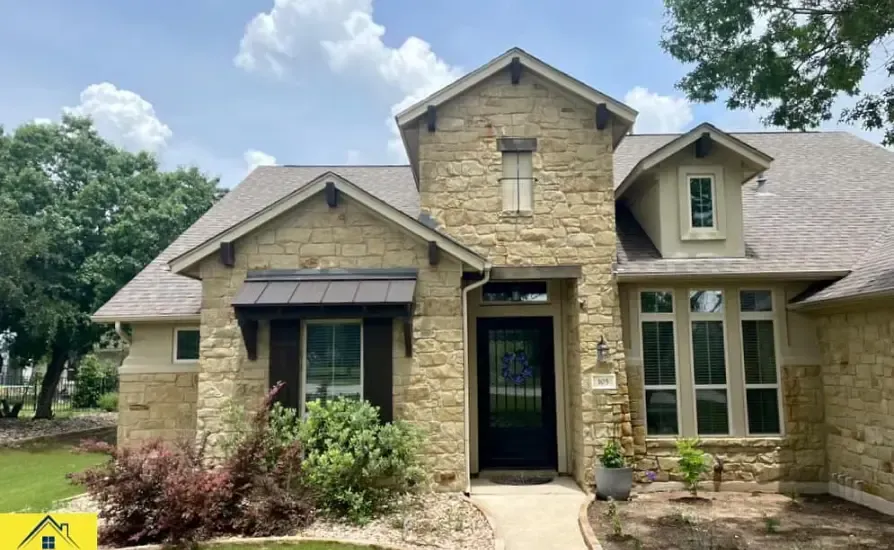 Beige stone house with dark front door, awning, and multiple windows under a blue sky.