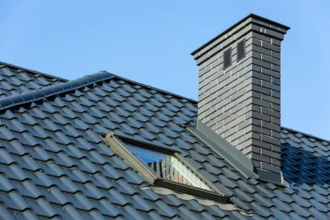 Dark gray tiled roof with chimney and skylight against a blue sky.