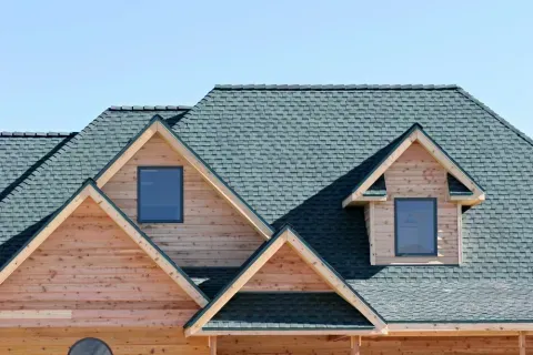 Wooden house under construction with green shingled roof, dormers, and blue sky.