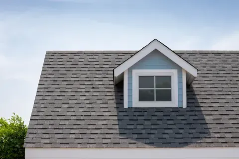 Gray shingled roof with a blue dormer window against a blue sky.