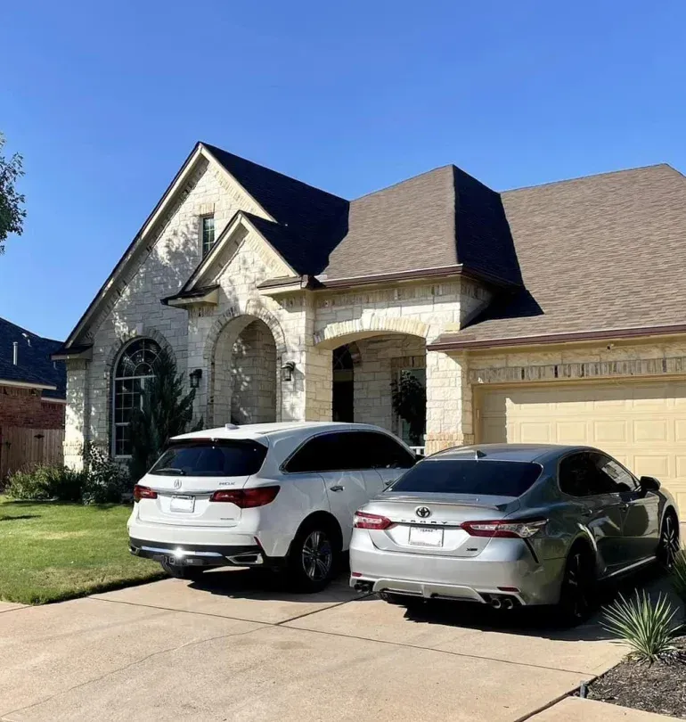 Two cars parked in a driveway in front of a stone-faced house on a sunny day.