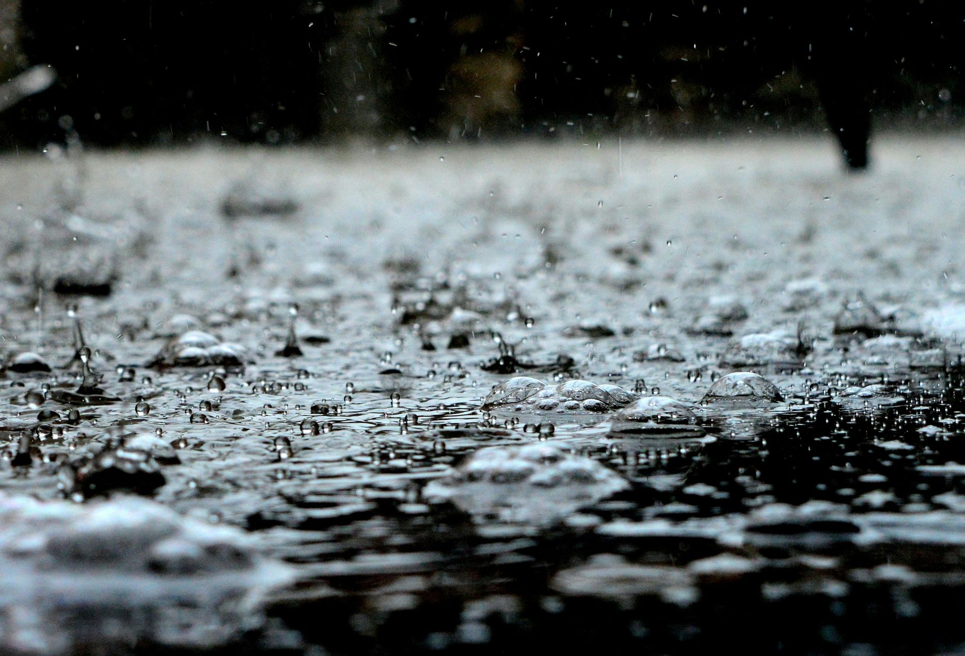 Raindrops hitting a puddle, creating ripples and splashes on a gray surface; blurry background.