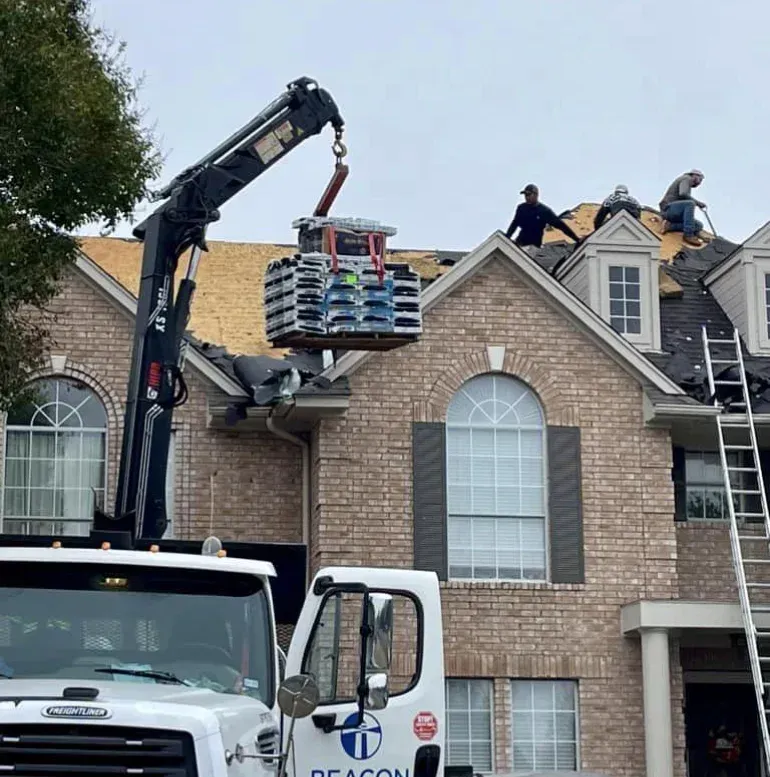Crane lifting roofing materials to workers on a house roof. White truck below.