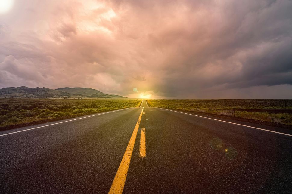 A road going through a field with the sun shining through the clouds.