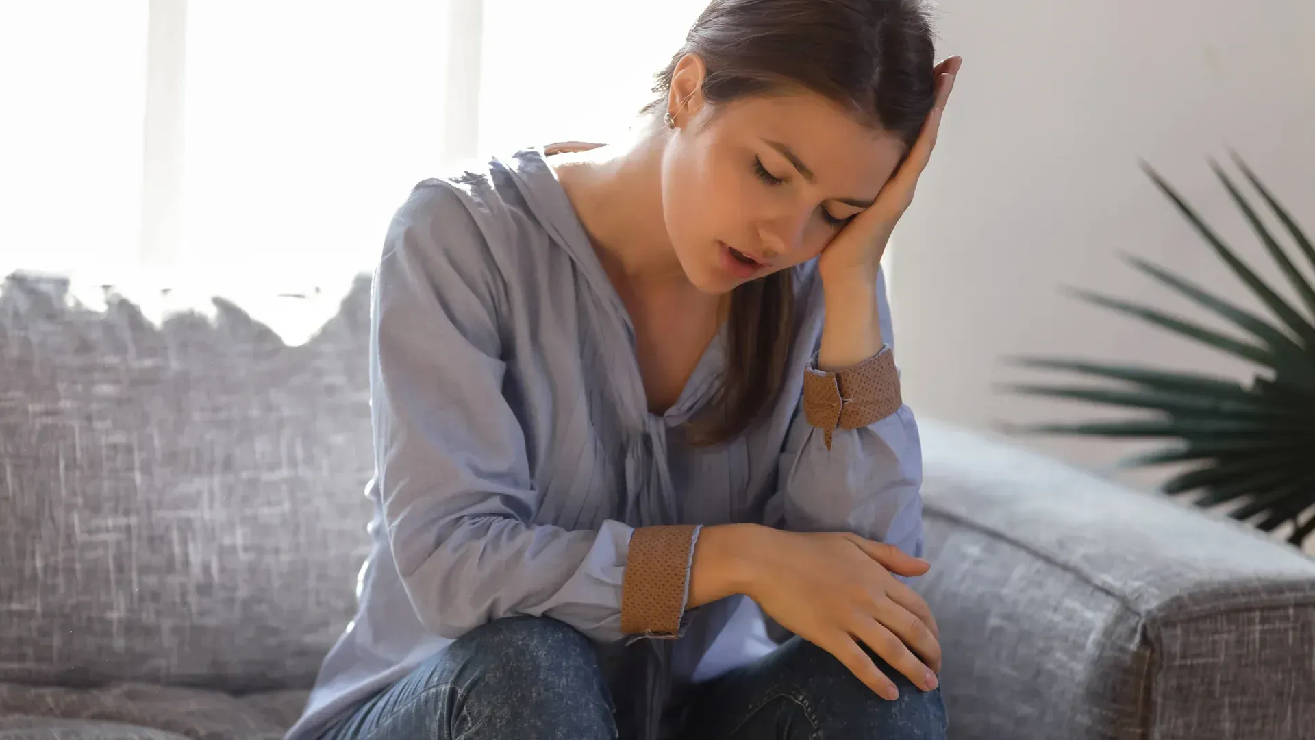 Woman with head in hand, looking down, seated on a couch; expressing sadness or exhaustion.