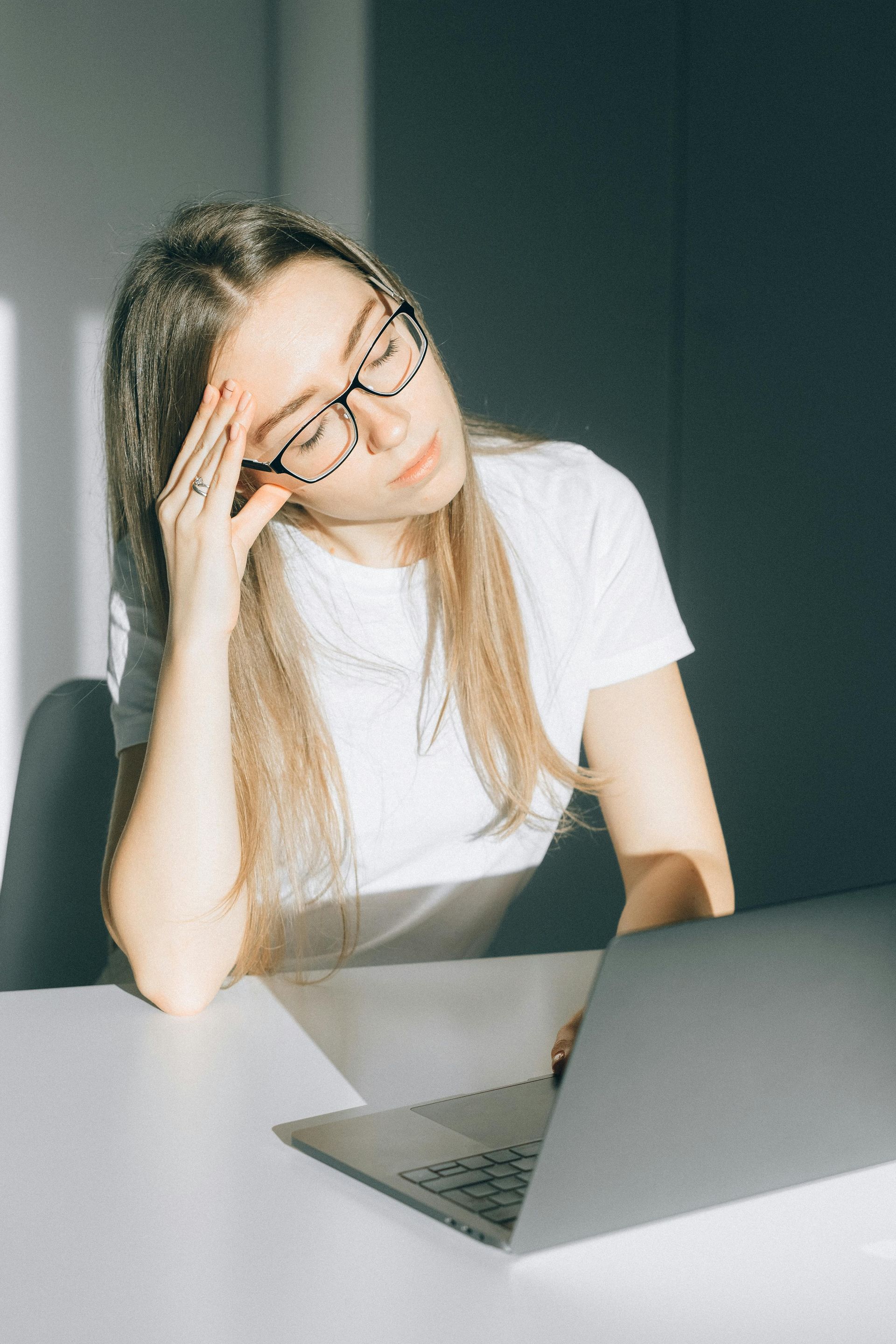 Woman with glasses and a white shirt looks stressed, hand on forehead, by a laptop.