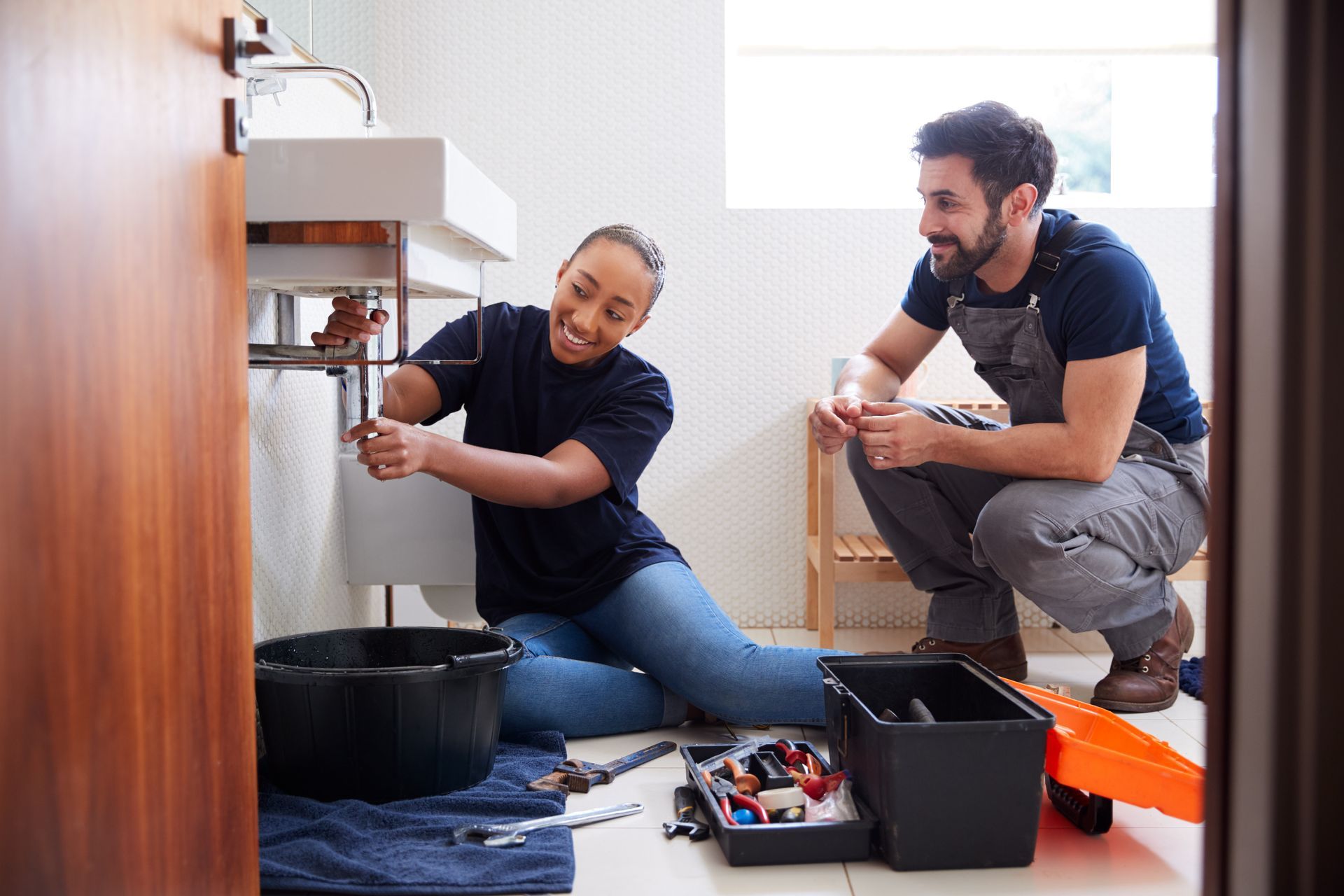 Two people repair a sink under a bathroom counter, with tools and a bucket on the floor. Two people repair a sink under a bathroom counter, with tools and a bucket on the floor.