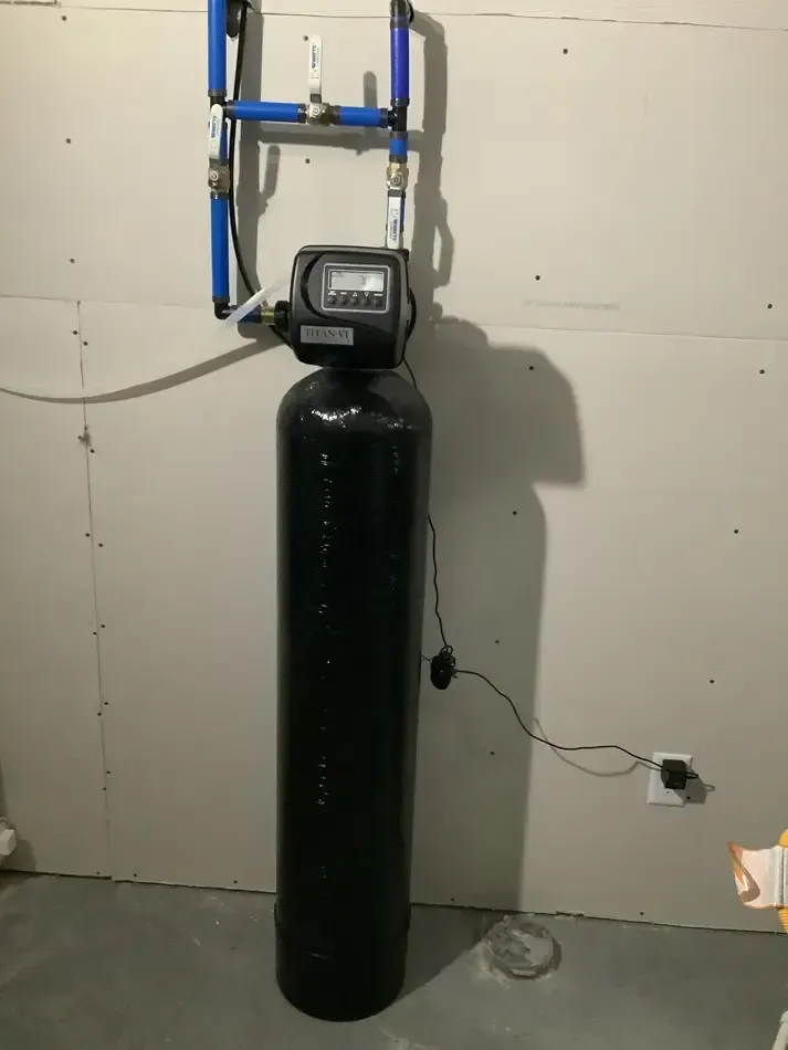 Black water softener tank with control valve in a utility room, mounted pipes above and a power cord beside it Black water softener tank with control valve in a utility room, mounted pipes above and a power cord beside it