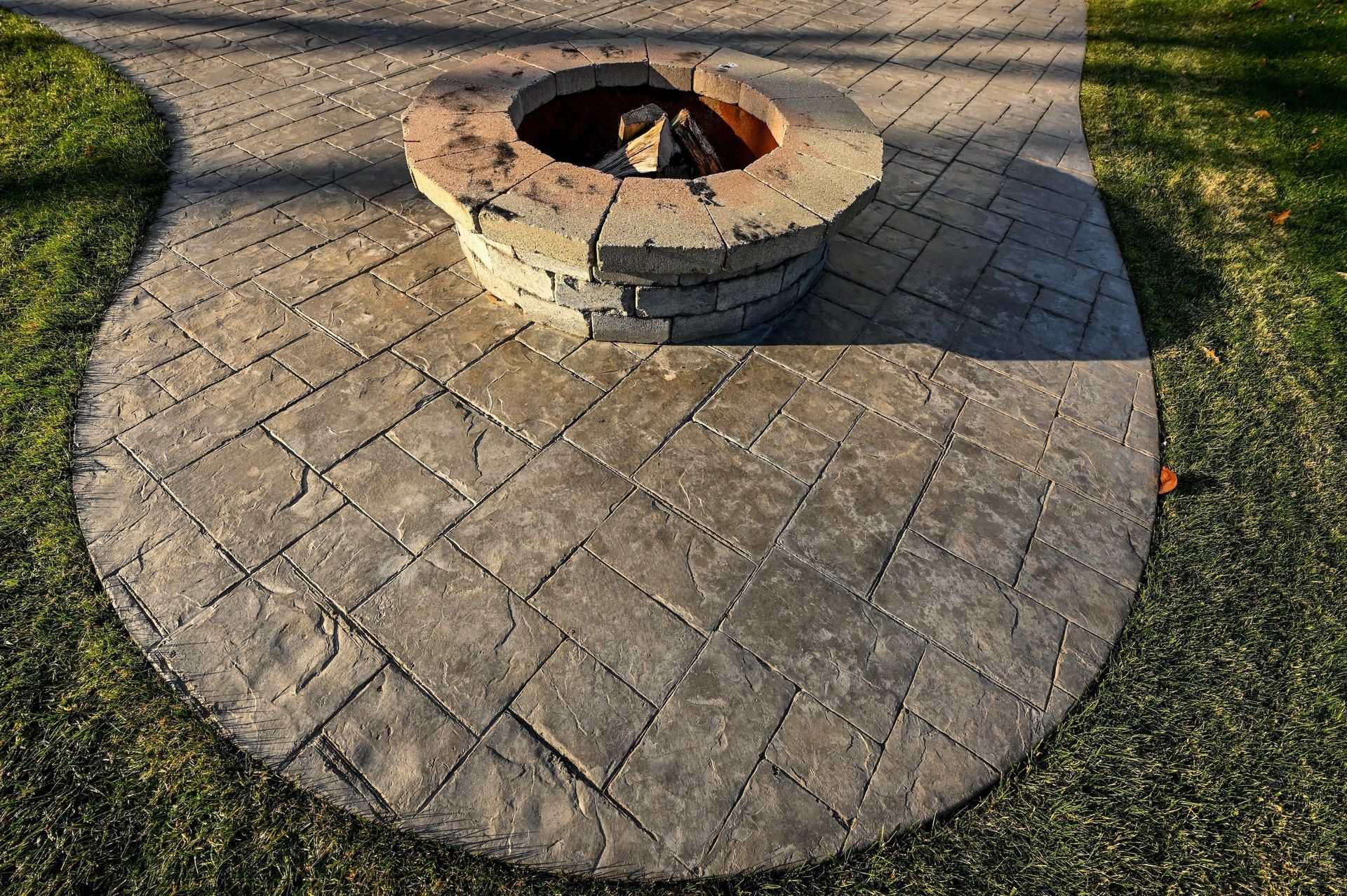Circular stone fire pit on a patterned gray patio, set in a grassy yard.