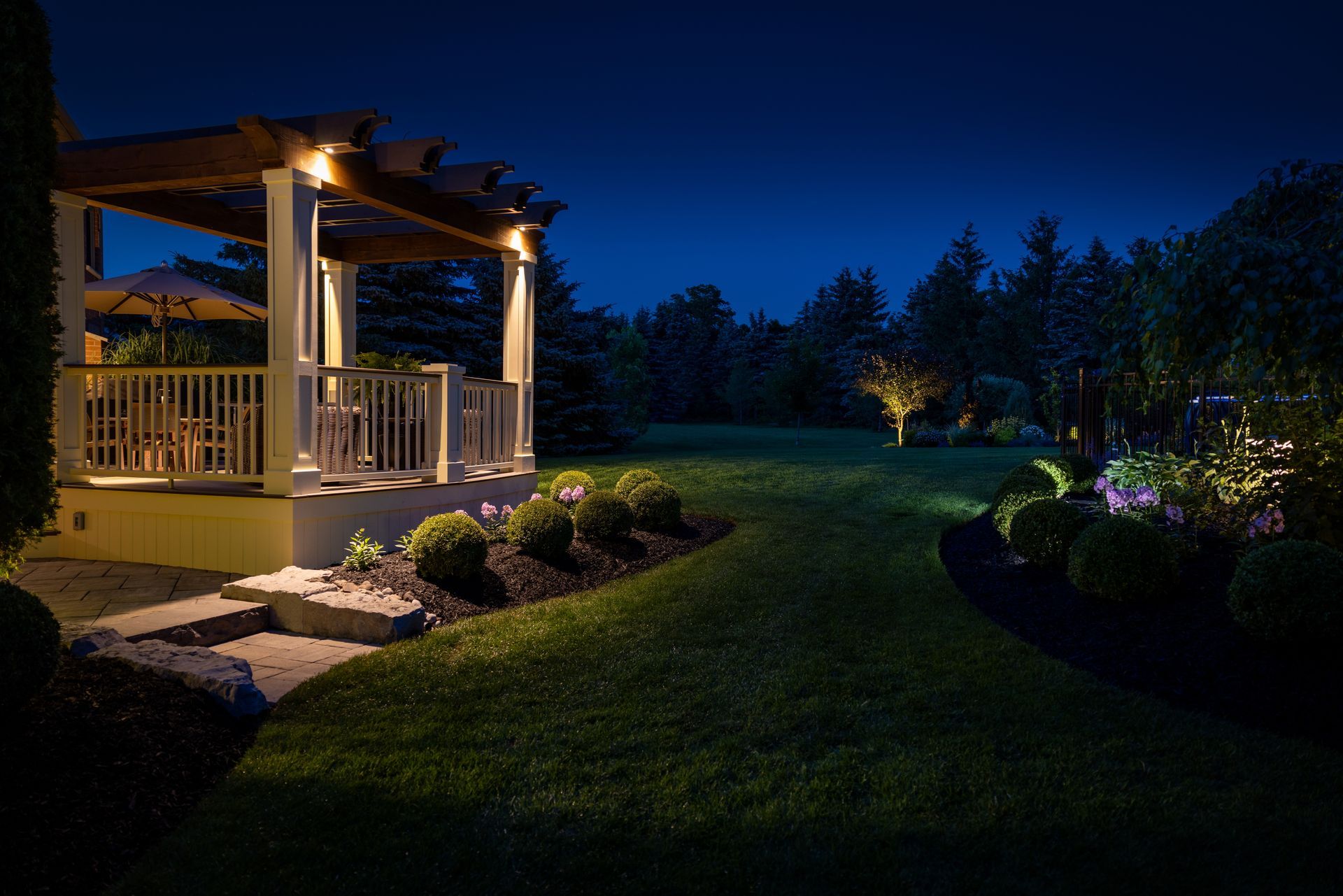 Night view of a lit gazebo on a lawn, surrounded by trees and illuminated landscaping lights.