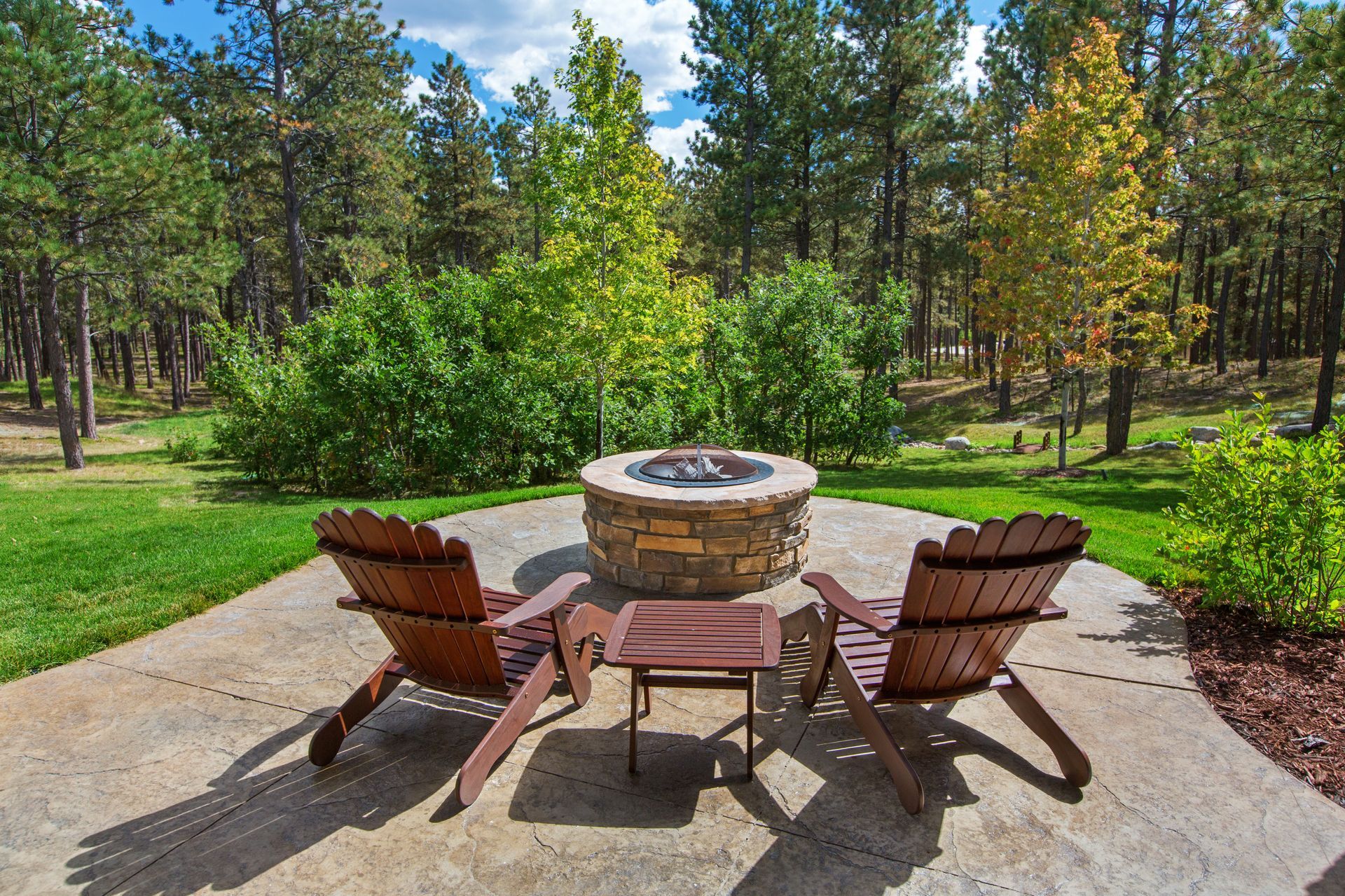 Two wooden chairs face a fire pit on a patio, surrounded by trees and greenery.