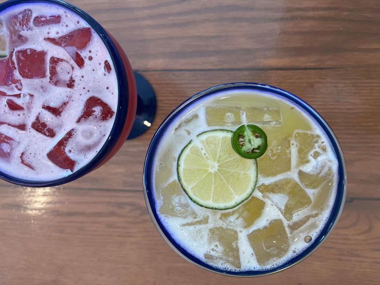 Two drinks are on a wooden table and one has a jalapeno on it