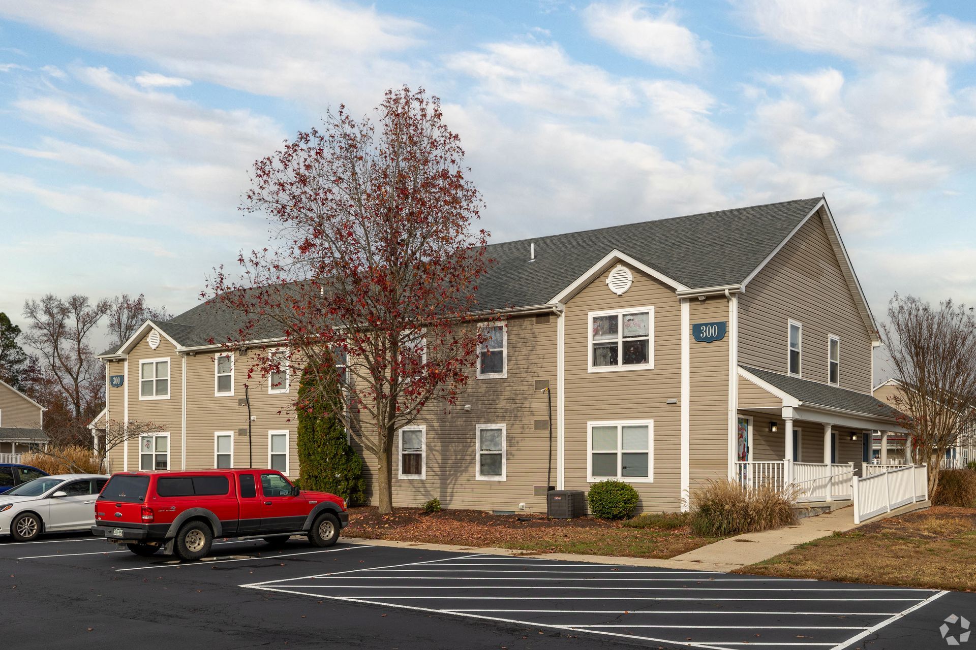 Two-story tan building with a red truck parked in the foreground. Blue sky and parking lot.