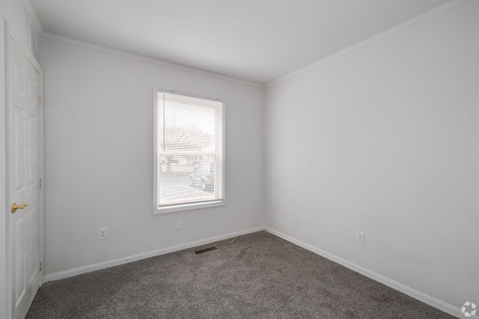 Empty bedroom with gray carpet, white walls, small window with blinds, and a closed white door.