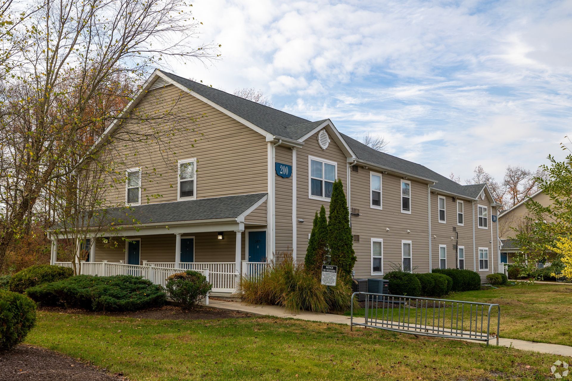 Beige apartment building with a porch and bike rack on a grassy area under a cloudy sky.