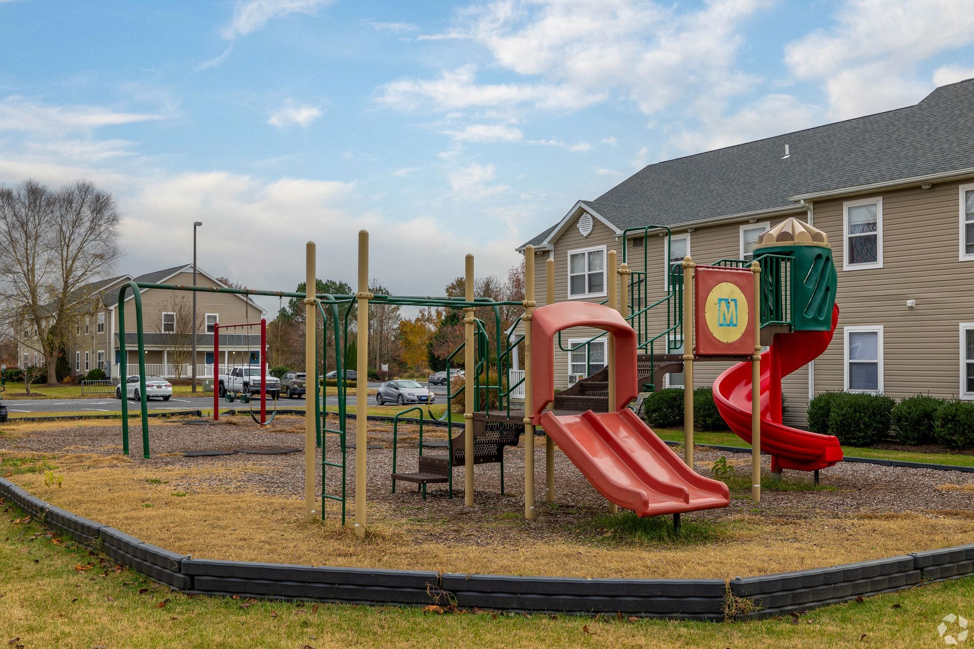 Playground with red and green slides and swings in front of an apartment building on a cloudy day.
