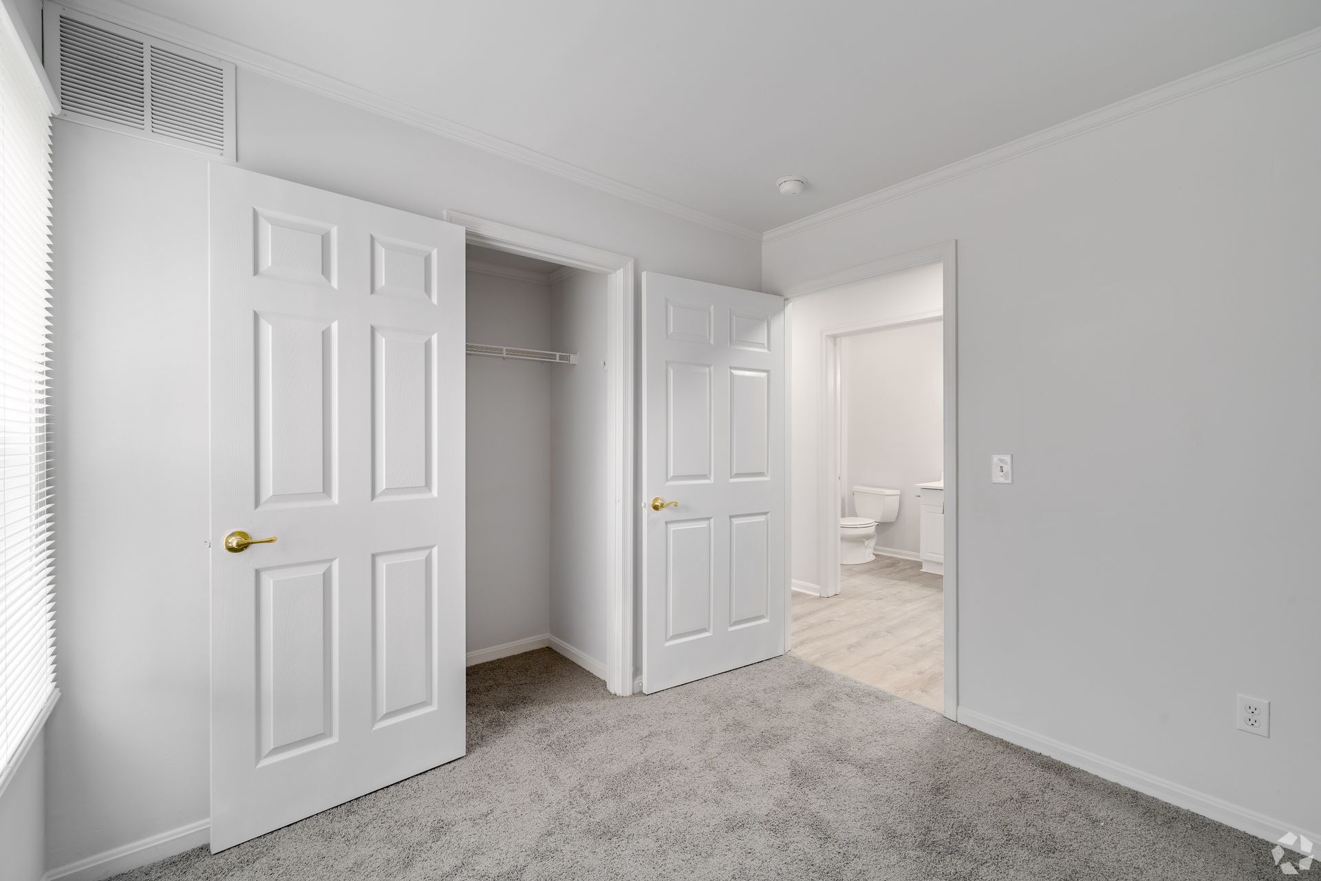 Empty bedroom with open closet and doorway to a bathroom. White walls, doors, and light gray carpet.
