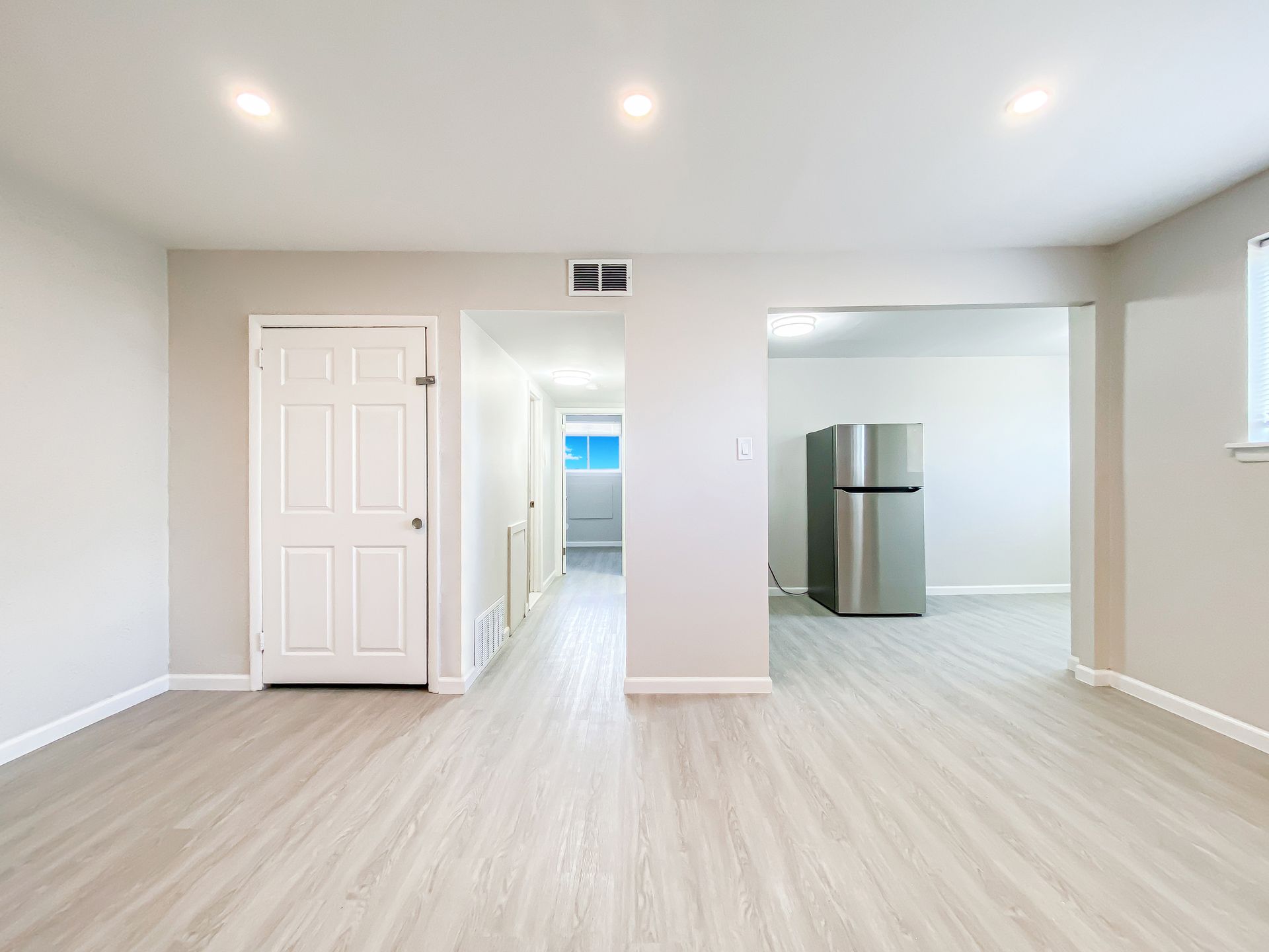 Empty interior with light wood-look flooring, white walls, and a silver refrigerator in the kitchen area.