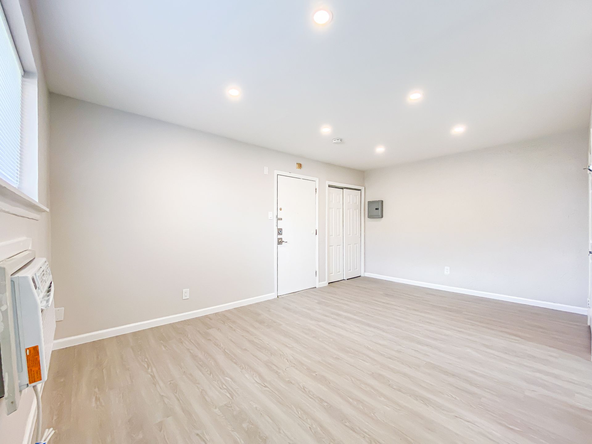 Empty room with light wood floors, white walls, recessed lighting, and a closed door.