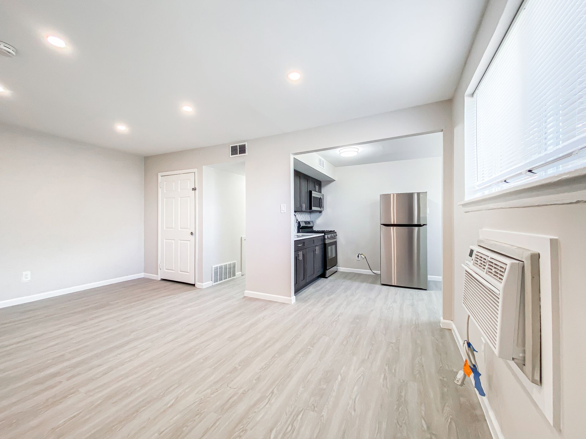 Empty living space with light wood-look flooring, leading to a dark kitchen with stainless steel appliances.