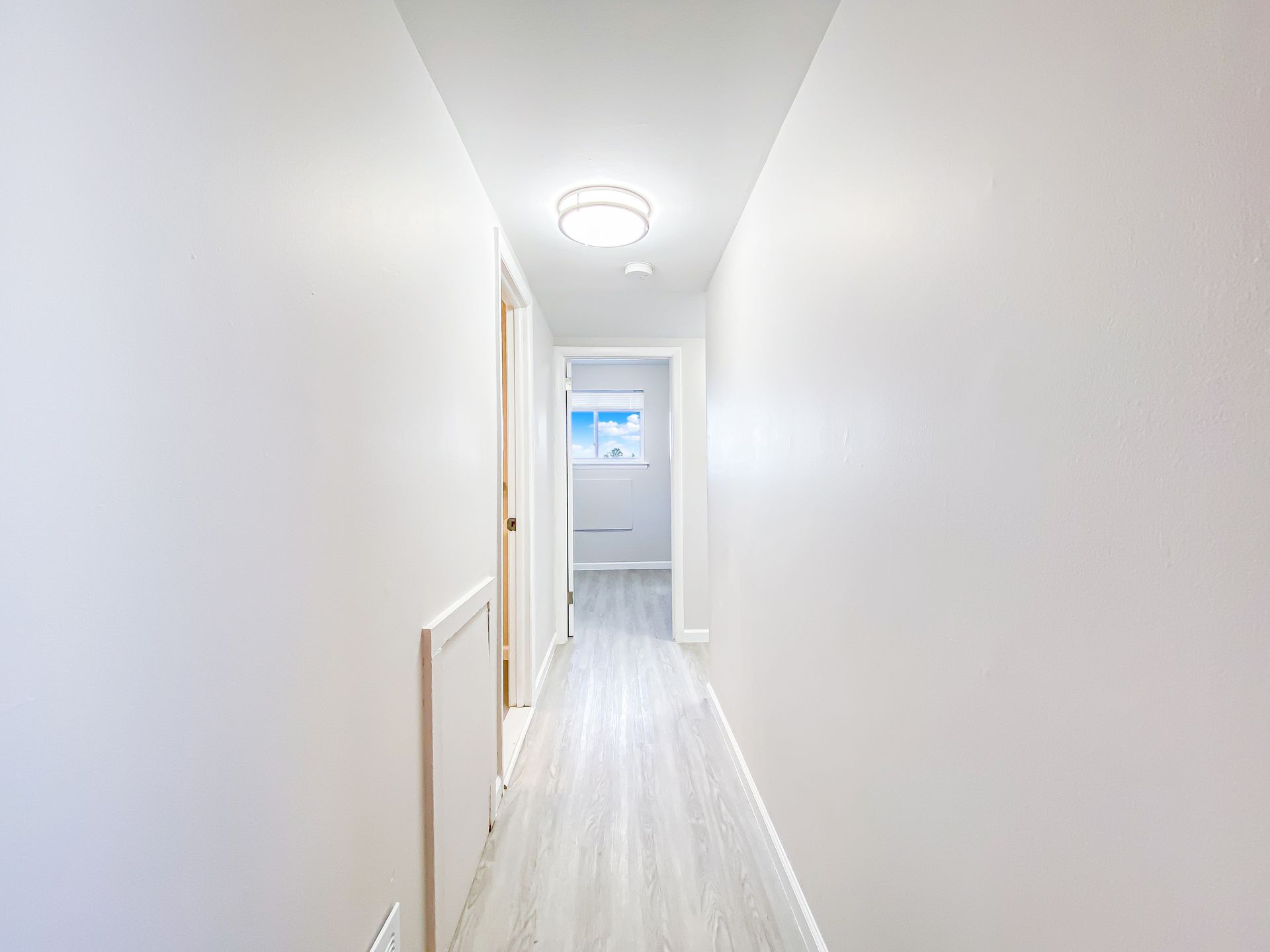 Narrow, bright hallway with light wood-look flooring and white walls, door at the end.