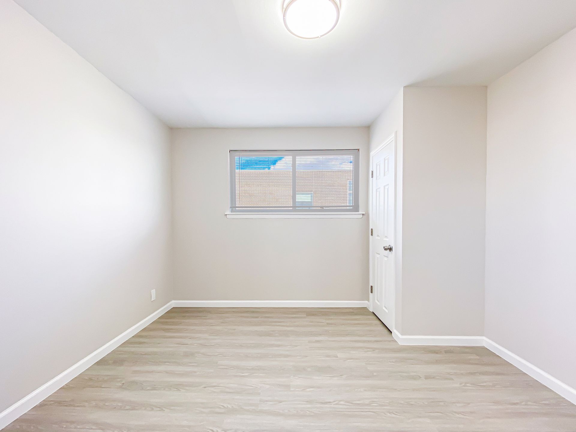 Empty, light-colored room with a window, door, and wooden floor.
