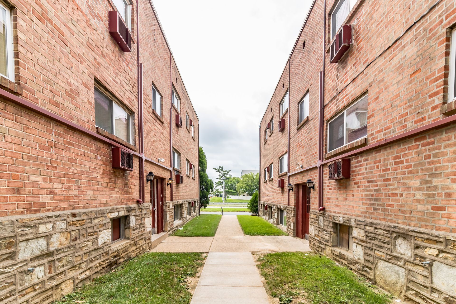 Two red brick apartment buildings face each other, with a concrete walkway and green space in between.