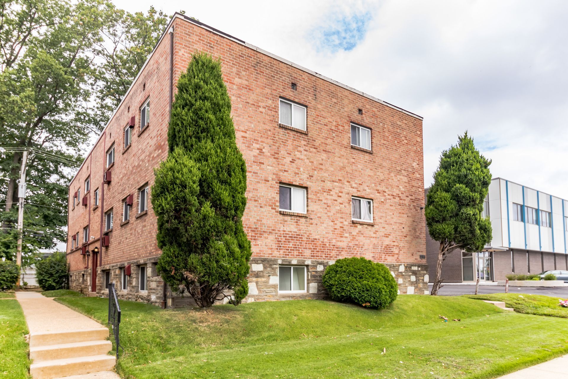 Three-story brick apartment building with stone foundation; green lawn and shrubs in front.