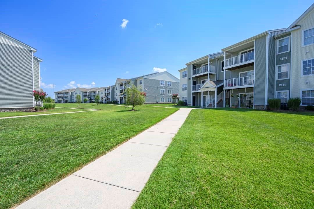 Pathway through a grassy lawn leading to apartment buildings under a bright blue sky.