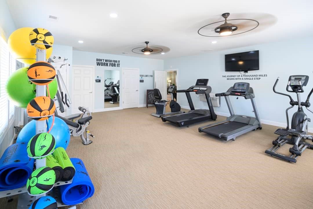 Gym with cardio machines, exercise balls, and weight rack, on beige carpet, light blue walls.