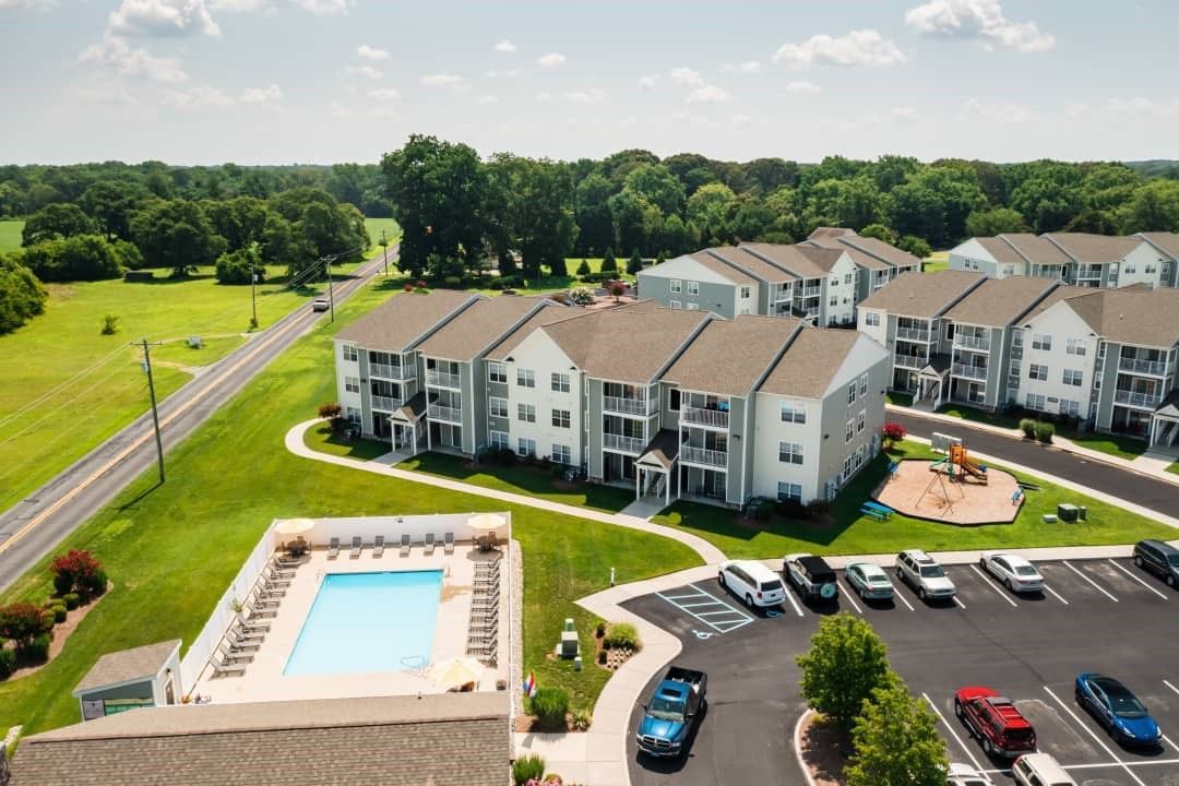 Aerial view of an apartment complex with a pool, parking, and a playground, surrounded by green grass and trees.