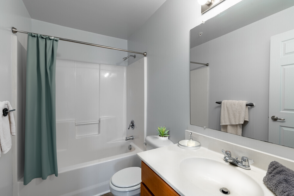 Bathroom with white tub/shower, toilet, vanity, and large mirror. Light blue walls and green shower curtain.