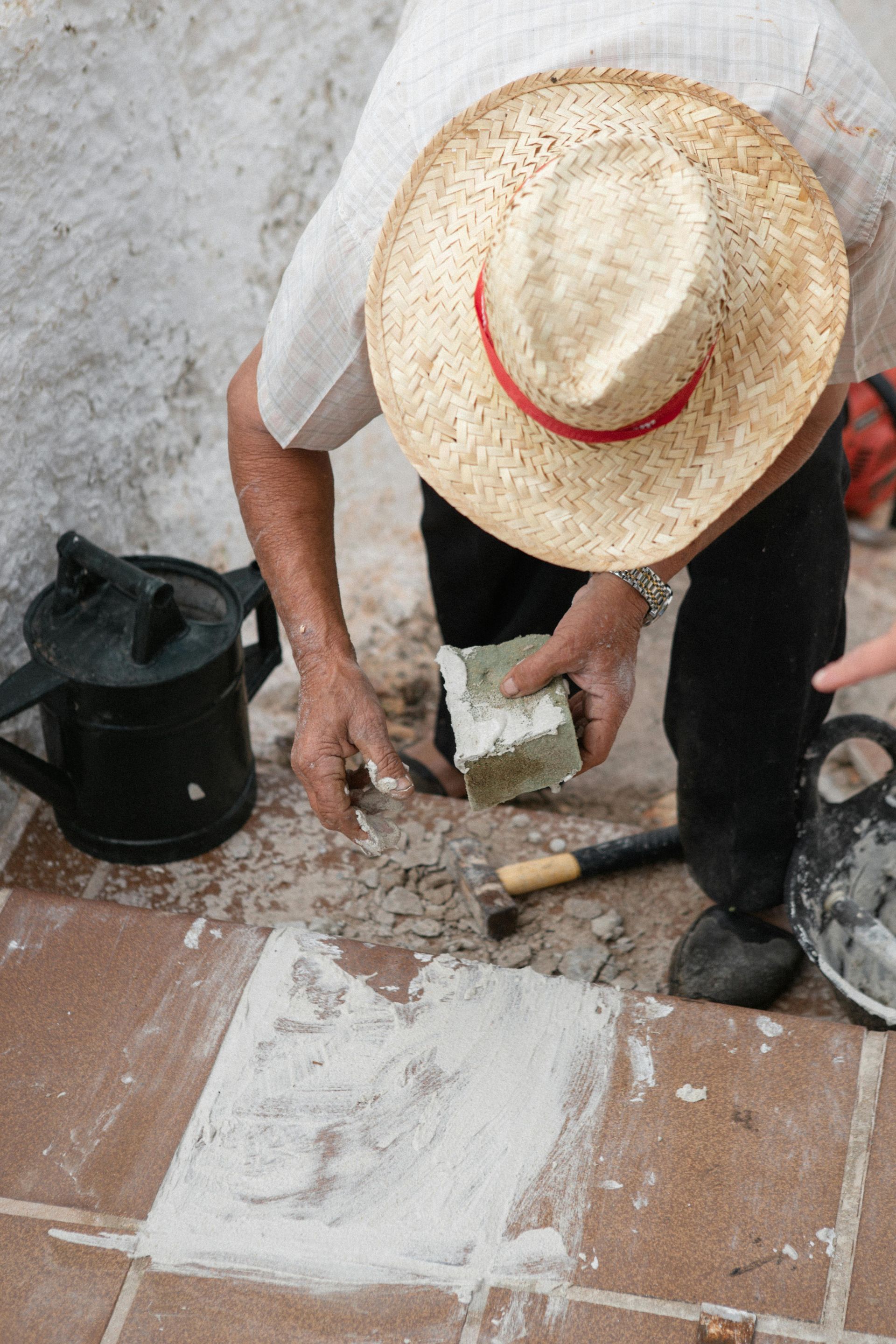 A person in a straw hat works on a tiled floor, holding a small stone block with mortar nearby.