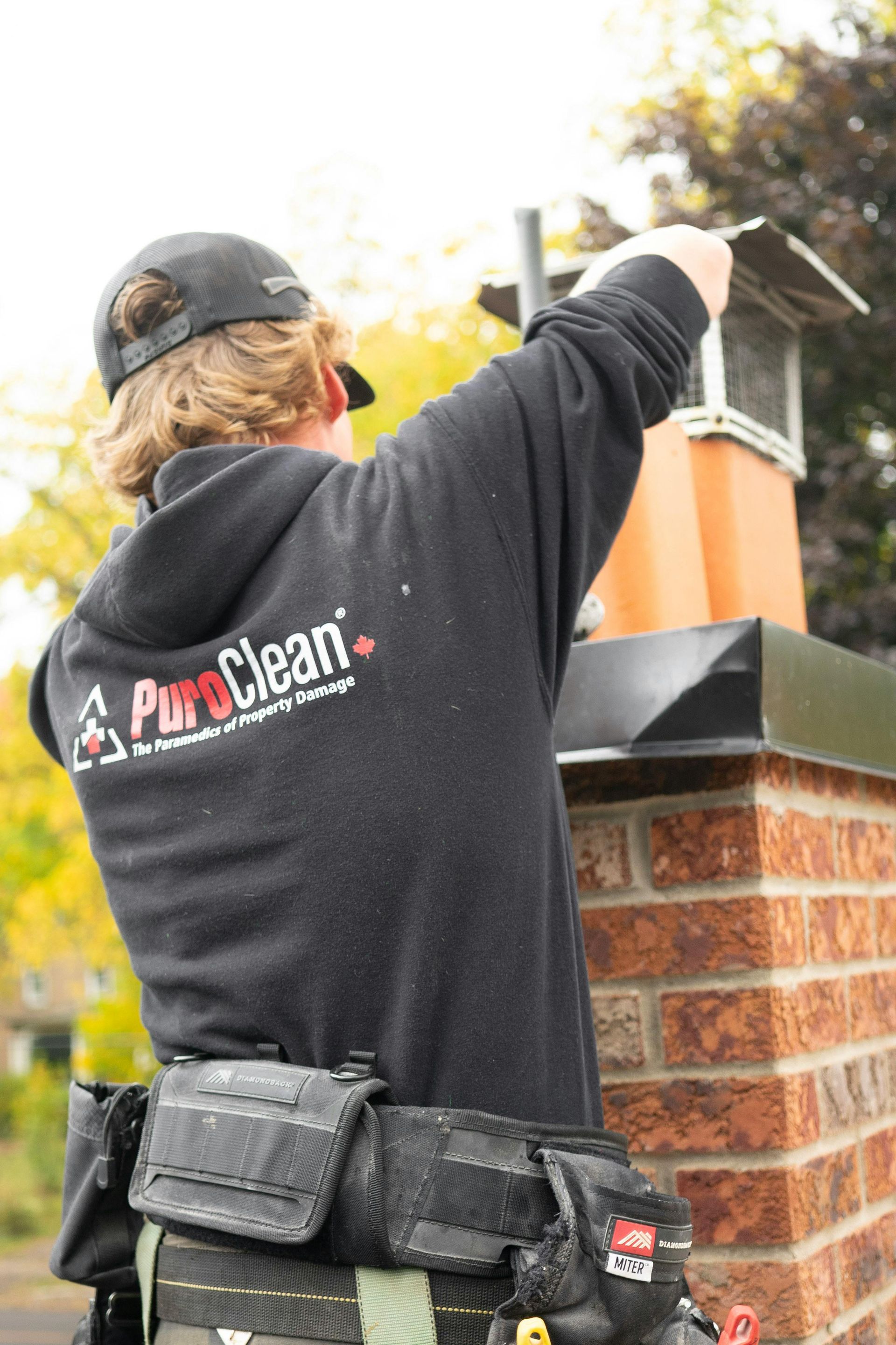 A person wearing a PuroClean hoodie and tool belt inspects a brick chimney outdoors.