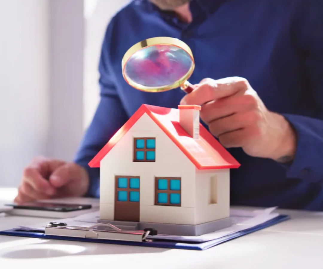 Person holding a magnifying glass over a small house model on a desk