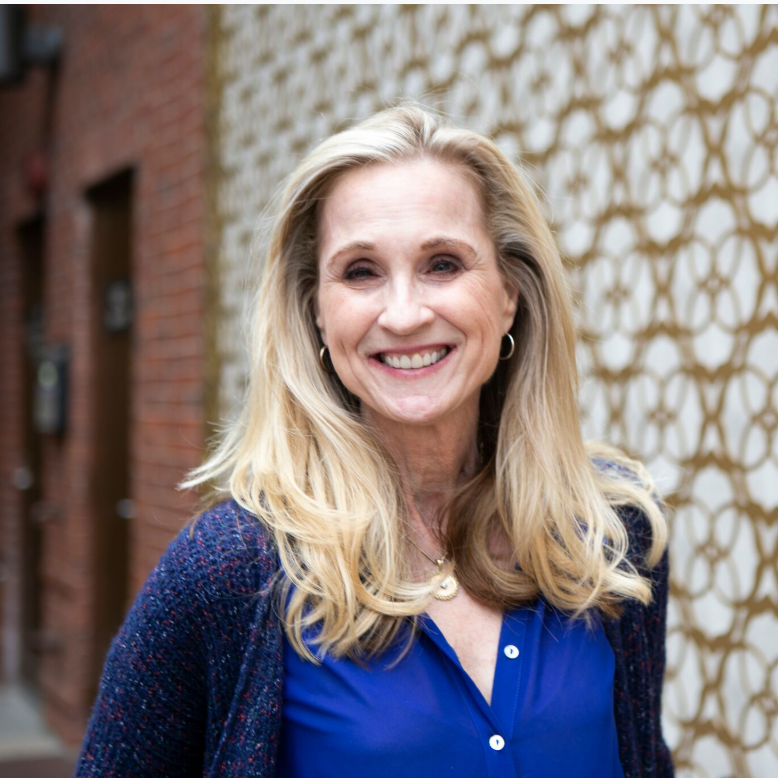 A woman in a blue shirt and blue cardigan is smiling in front of a brick wall.