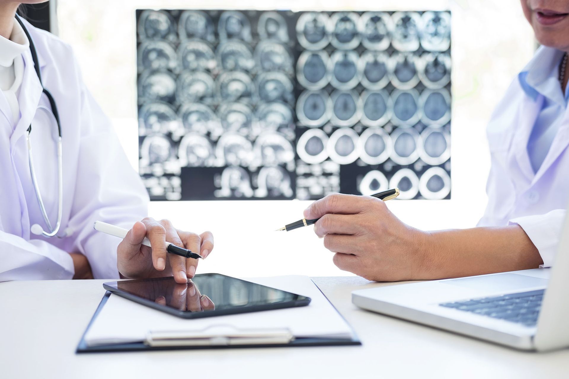 A doctor discusses with another doctor using a tablet the results of a brain x-ray in the background. A doctor discusses with another doctor using a tablet the results of a brain x-ray in the background.