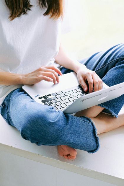 A person in a white t-shirt and blue jeans sits cross-legged while using a silver laptop.