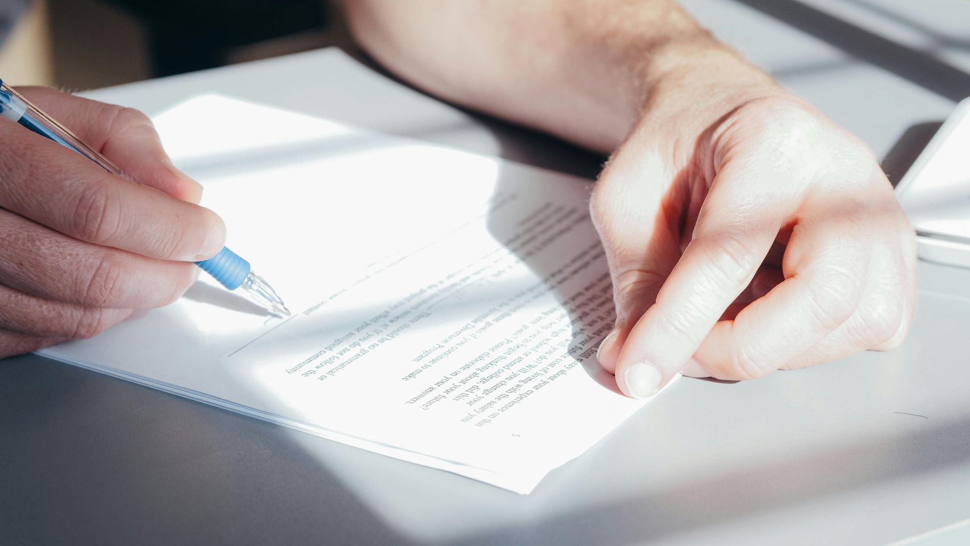 A close-up view of hands holding a blue pen and writing on a white document under bright, natural light.